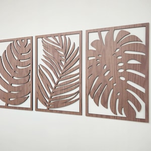 May include: Three wooden wall decor pieces featuring cutout designs of tropical leaves. The leaves are in a light brown color and are arranged in a horizontal row.