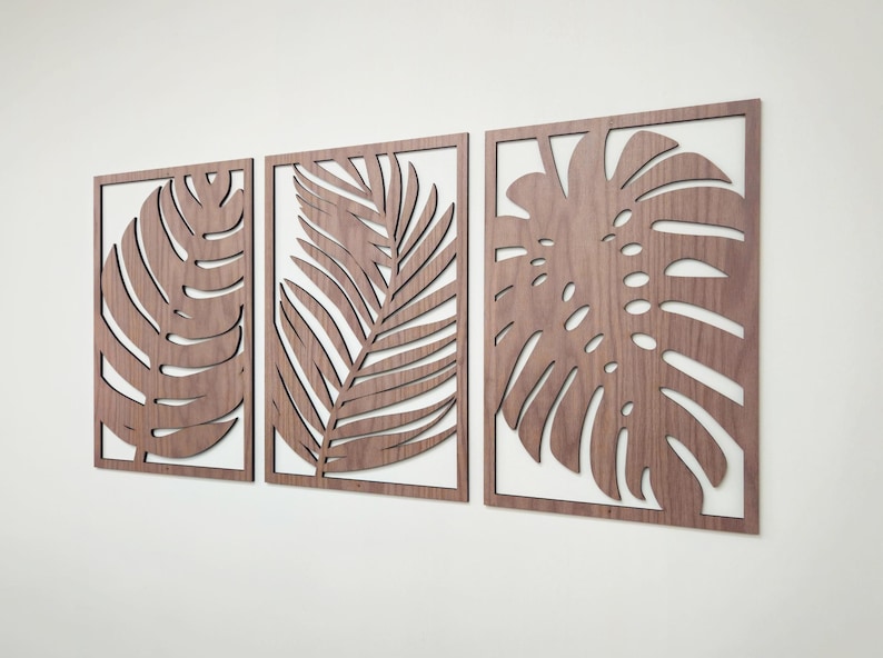 May include: Three wooden wall decor pieces featuring cutout designs of tropical leaves. The leaves are in a light brown color and are arranged in a horizontal row.