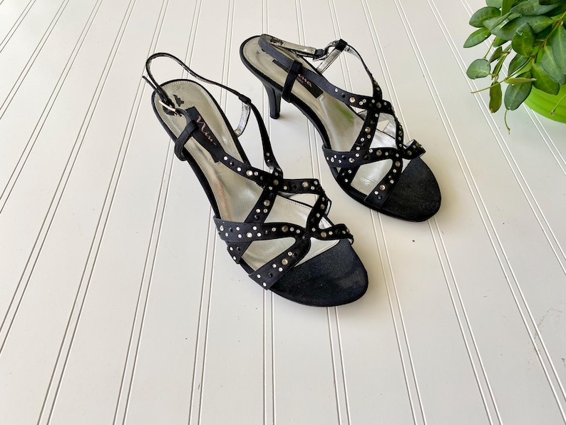 K&ouml;nnte beinhalten: Schwarze Riemchensandalen mit silbernen Nieten und einem Stiletto-Absatz. Die Sandalen haben mehrere Riemen, die sich &uuml;ber Fu&szlig; und Kn&ouml;chel kreuzen. Das Innere des Schuhs ist silberfarben.