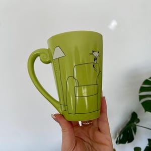 May include: A lime green ceramic mug with a large handle. The mug features a black and white illustration of a cat sitting on a couch next to a lamp. The mug is held by a person.