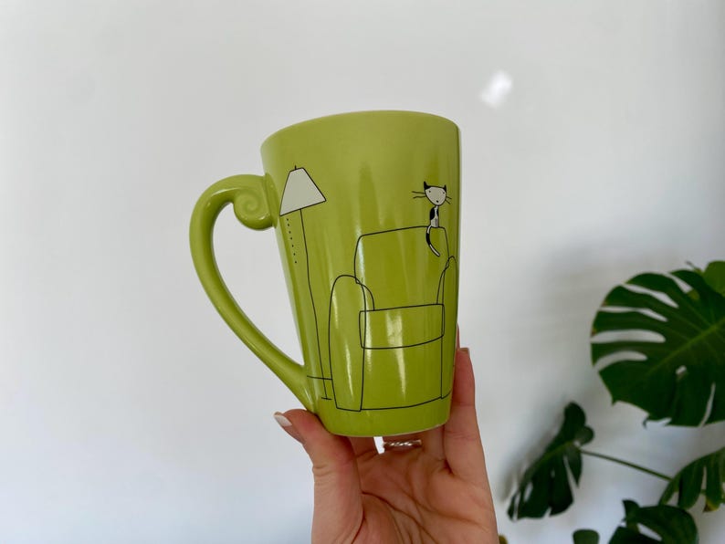 May include: A lime green ceramic mug with a large handle. The mug features a black and white illustration of a cat sitting on a couch next to a lamp. The mug is held by a person.