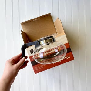 May include: A clear glass coffee decanter with a black handle and a silver lid, inside a cardboard box. The box has a red and white label with the words "coffee decanter" and "easy pour".