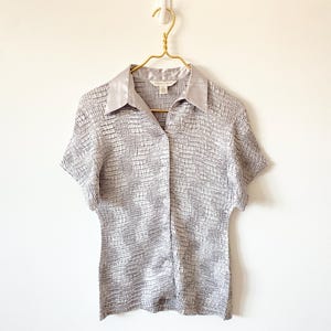 Op de afbeelding: Een lichtgrijs shirt met korte mouwen en een textuurpatroon in de vorm van een raster. Het shirt heeft een kraag en is dichtgeknoopt. Het hangt aan een goudkleurige hanger. Het shirt is gemaakt van licht materiaal.
