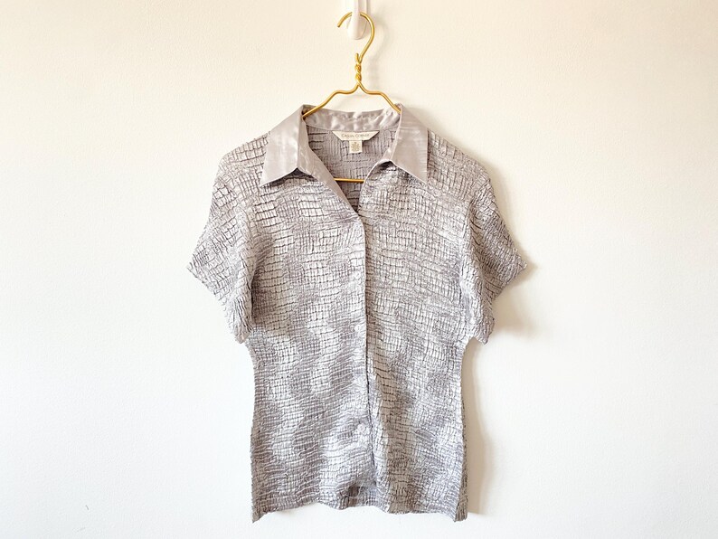 Op de afbeelding: Een lichtgrijs shirt met korte mouwen en een textuurpatroon in de vorm van een raster. Het shirt heeft een kraag en is dichtgeknoopt. Het hangt aan een goudkleurige hanger. Het shirt is gemaakt van licht materiaal.
