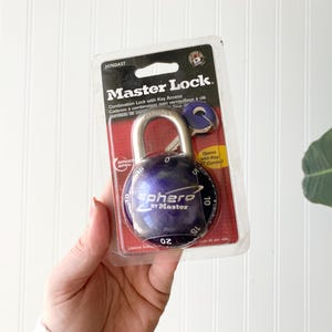May include: A Master Lock combination lock with key access, in a clear plastic package. The lock is dark blue with a silver shackle and a combination dial. The packaging includes the Master Lock logo and product information.