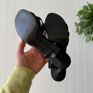 K&ouml;nnte beinhalten: Ein Paar schwarze Sandalen mit hohen Abs&auml;tzen, d&uuml;nnen Riemen und einem kleinen Absatz. Die Sohlen weisen Gebrauchsspuren auf. Die Sandalen werden in der Hand einer Person gehalten.