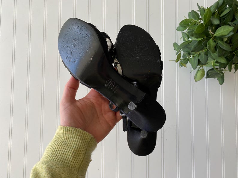 K&ouml;nnte beinhalten: Ein Paar schwarze Sandalen mit hohen Abs&auml;tzen, d&uuml;nnen Riemen und einem kleinen Absatz. Die Sohlen weisen Gebrauchsspuren auf. Die Sandalen werden in der Hand einer Person gehalten.