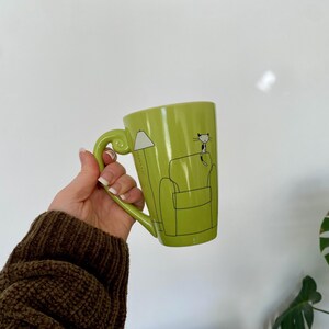 May include: A lime green ceramic mug with a large handle. The mug features a black and white illustration of a cat sitting on a couch next to a lamp. The mug is held in a person's hand.