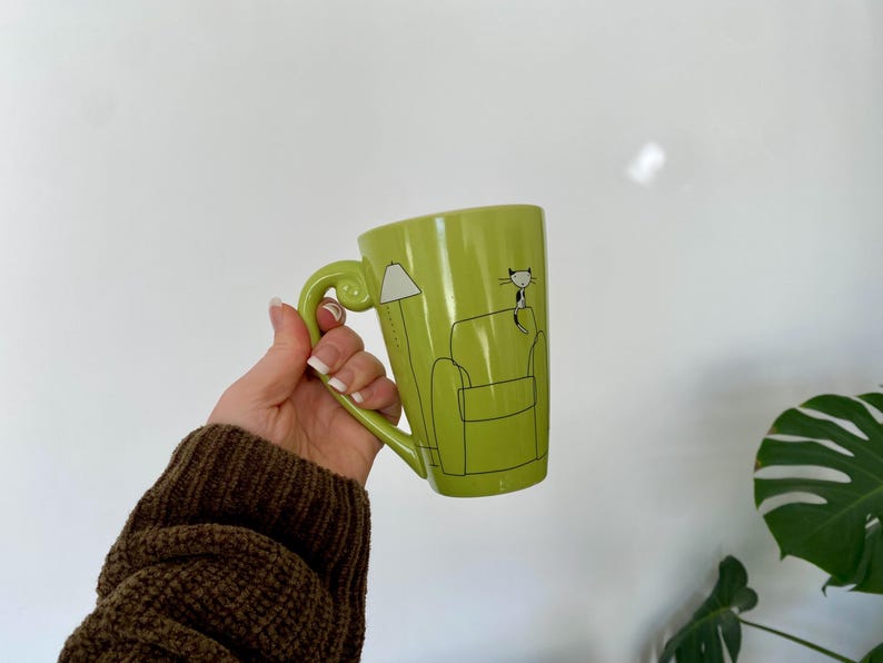 May include: A lime green ceramic mug with a large handle. The mug features a black and white illustration of a cat sitting on a couch next to a lamp. The mug is held in a person's hand.