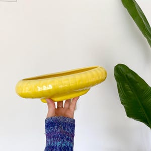 May include: A bright yellow ceramic bowl with a wide, shallow basin and a distinctively ribbed, scalloped outer rim. The bowl features a glossy, reflective finish that catches the light, highlighting its smooth, curved edges and circular form.