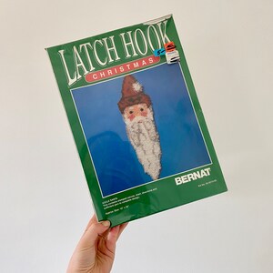 May include: A green and white latch hook kit featuring a Santa Claus design. The kit includes stamped canvas, chart, directions and yarn. Approximate size 16" x 20".