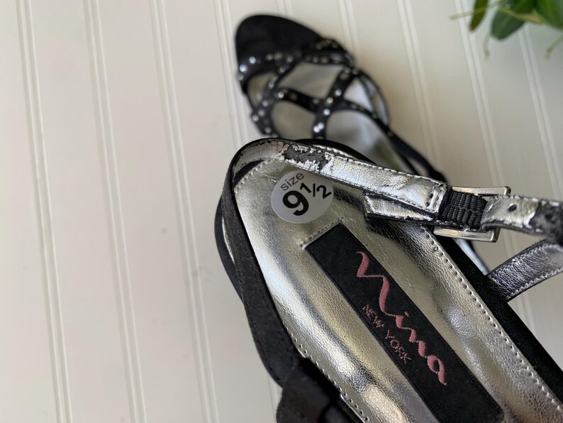 K&ouml;nnte beinhalten: Schwarze Riemchen-High Heels mit silbernen Akzenten und einem Gr&ouml;&szlig;enetikett 9 1/2. Die Schuhe haben eine spitze Zehenpartie und einen Schnallenverschluss. Der Markenname "Nina New York" ist auf der Innensohle sichtbar.