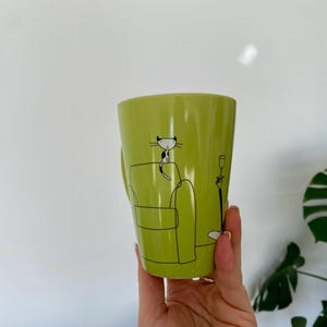 May include: A lime green ceramic mug with a black and white cat illustration. The cat is sitting on a green couch with a lamp and a wine glass. The mug has a handle on the side.