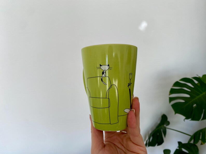 May include: A lime green ceramic mug with a black and white cat illustration. The cat is sitting on a green couch with a lamp and a wine glass. The mug has a handle on the side.