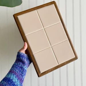 May include: A rectangular wooden tray with six cream-colored tiles. The tray has a dark brown wooden frame. The tiles are arranged in two rows of three. The tray is held by a person wearing a blue and purple sweater.