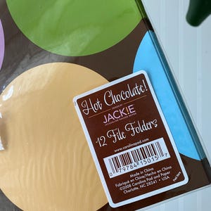 May include: A pack of 12 file folders featuring a brown background with large green, light blue, and tan circles. A white label on the front reads Hot Chocolate! Design by JACK!E Jacqueline Savage McFee 12 File Folders with a barcode and text.