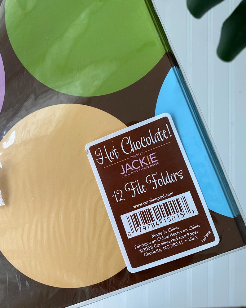 May include: A pack of 12 file folders featuring a brown background with large green, light blue, and tan circles. A white label on the front reads Hot Chocolate! Design by JACK!E Jacqueline Savage McFee 12 File Folders with a barcode and text.