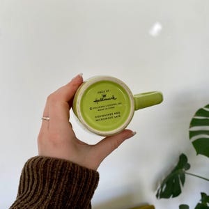 May include: A green Hallmark mug with a matching handle. The bottom of the mug is visible, displaying the Hallmark logo and text indicating it is dishwasher and microwave safe. The mug is held against a white background.