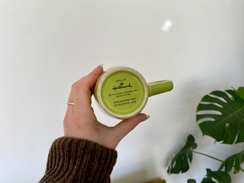 May include: A green Hallmark mug with a matching handle. The bottom of the mug is visible, displaying the Hallmark logo and text indicating it is dishwasher and microwave safe. The mug is held against a white background.