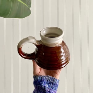 May include: A handmade ceramic mug with a brown exterior and a white interior. The handle is white and brown. The mug is held up by a person. The mug is approximately 4 inches tall.
