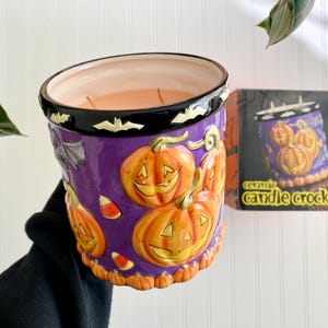 May include: A ceramic candle crock with a purple base, orange pumpkins, and a black rim with white bats. The candle crock has a decorative design and the words "ceramic candle crock" on the packaging.