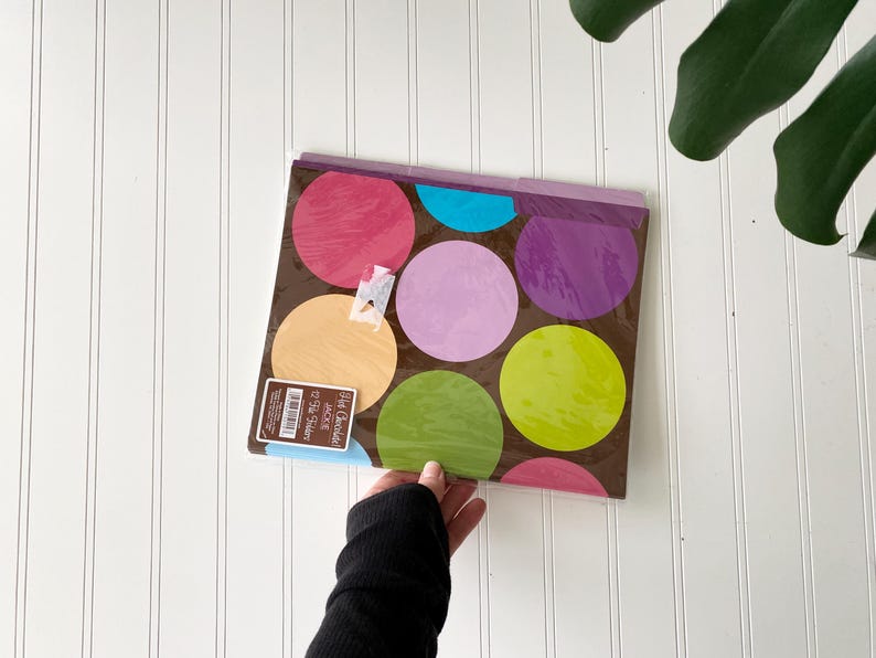 May include: A set of 12 file folders with a brown background featuring large, colorful polka dots in shades of pink, purple, light blue, yellow, and lime green. The packaging includes a label with the text Hot Chocolate Jackie 12 File Folders.