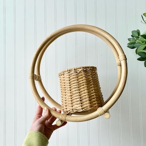 May include: A round, natural-toned rattan wall basket with a woven basket insert. The basket is held within a double-ring frame. The design is a light brown color and is suitable for displaying plants or other decorative items.
