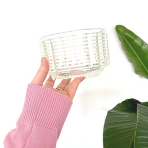 May include: A clear, vintage glass dish with a textured, woven-like pattern. The dish is held up against a white background, with a pink sweater sleeve in the frame. A green leaf is visible in the background.