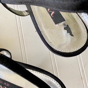 K&ouml;nnte beinhalten: Nahaufnahme von schwarzen Satin-High-Heel-Sandalen. Die Innensohle ist silberfarben mit einem schwarzen Etikett mit der Aufschrift "nina" in Pink. Die Sandalen haben einen d&uuml;nnen Kn&ouml;chelriemen und eine schwarze Sohle. Die Schuhe befinden sich auf einer wei&szlig;en Oberfl&auml;che.