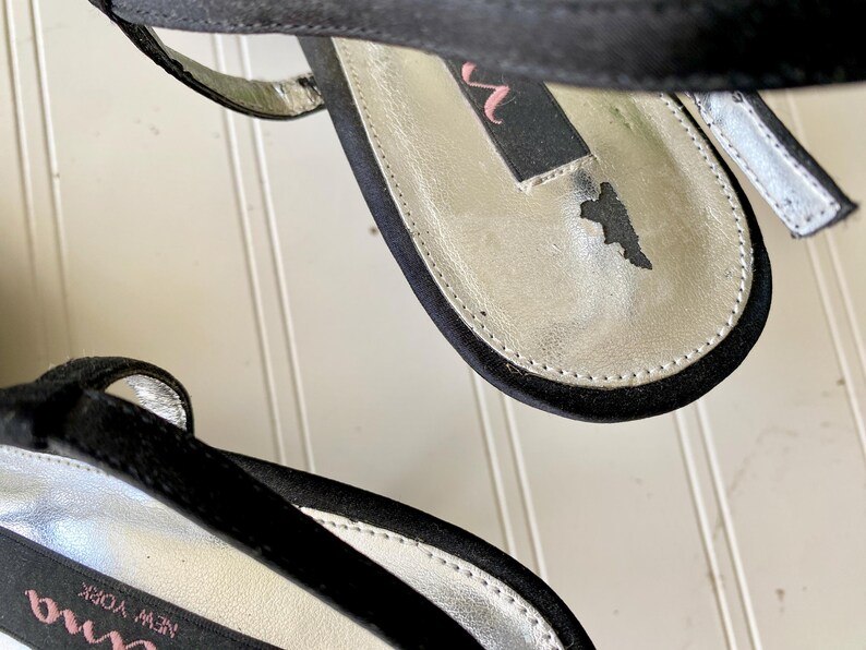 K&ouml;nnte beinhalten: Nahaufnahme von schwarzen Satin-High-Heel-Sandalen. Die Innensohle ist silberfarben mit einem schwarzen Etikett mit der Aufschrift "nina" in Pink. Die Sandalen haben einen d&uuml;nnen Kn&ouml;chelriemen und eine schwarze Sohle. Die Schuhe befinden sich auf einer wei&szlig;en Oberfl&auml;che.