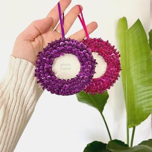 May include: Two circular hanging photo frames, one in deep purple and one in bright magenta, both covered in shimmering sequins. Each frame features a central circular opening with a white insert labeled photo frame and a matching ribbon loop.