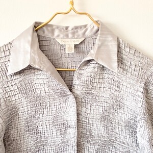 Op de afbeelding: Een zilverkleurige blouse met korte mouwen en een textuur met een rasterpatroon. De blouse heeft een satijnen kraag en hangt aan een goudkleurige hanger. Het label luidt "CASUAL CORNER".