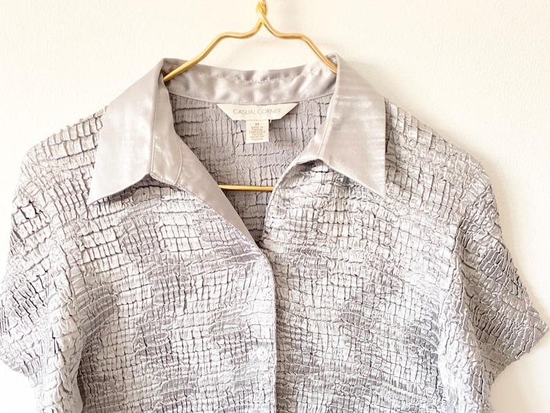 Op de afbeelding: Een zilverkleurige blouse met korte mouwen en een textuur met een rasterpatroon. De blouse heeft een satijnen kraag en hangt aan een goudkleurige hanger. Het label luidt "CASUAL CORNER".