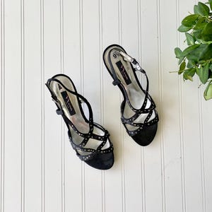 K&ouml;nnte beinhalten: Paar schwarze Sandalen mit hohen Abs&auml;tzen und Riemchen mit silberfarbenen Nieten. Die Sandalen haben einen Slingback-Riemen und einen Schnallenverschluss. Der Markenname "Nina" ist auf der Innensohle sichtbar. Gr&ouml;&szlig;e 9 1/2.