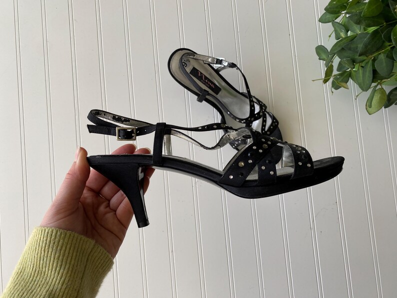 K&ouml;nnte beinhalten: Schwarze Satin-Sandalen mit kleinem Absatz. Die Sandalen haben mehrere Riemen mit silbernen Akzenten und kleinen, dekorativen Nieten. Der Kn&ouml;chelriemen hat einen Schnallenverschluss. Die Schuhe sind auf wei&szlig;em Hintergrund.
