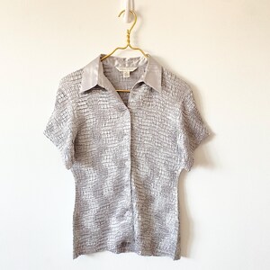 Op de afbeelding: Een shirt met korte mouwen in een textuur, zilver-grijze stof. Het shirt heeft een satijnen kraag en hangt aan een goudkleurige hanger tegen een witte muur. De stof heeft een subtiel, rasterachtig patroon.