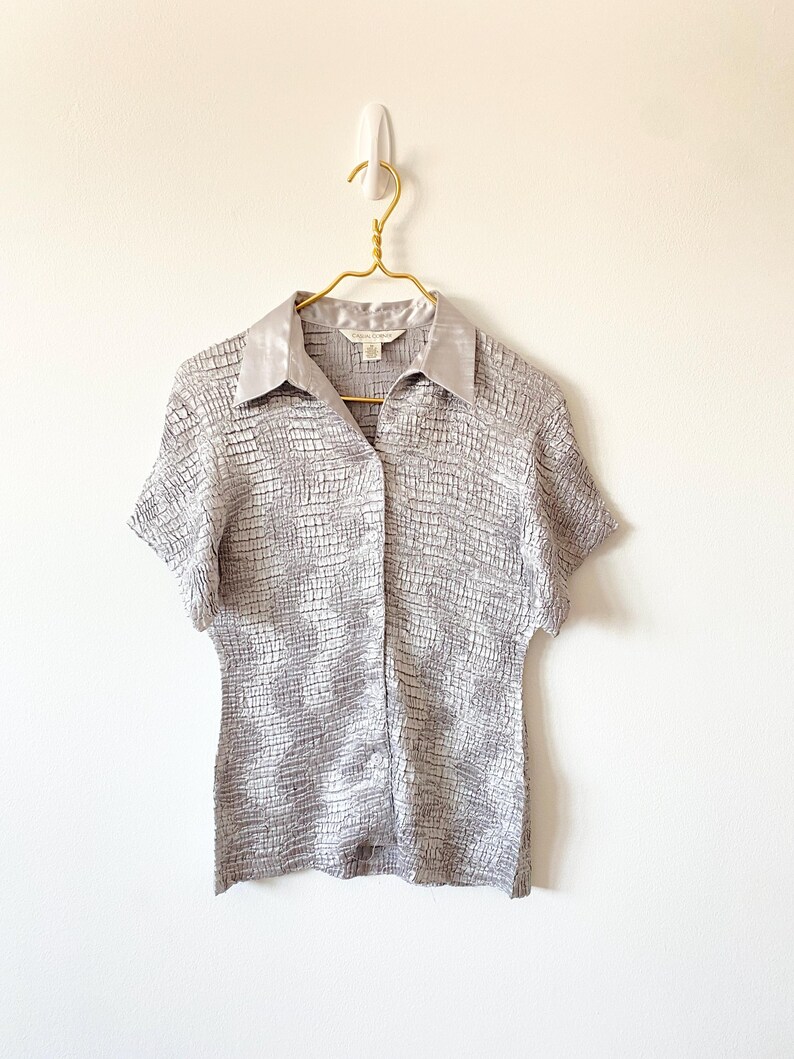 Op de afbeelding: Een shirt met korte mouwen in een textuur, zilver-grijze stof. Het shirt heeft een satijnen kraag en hangt aan een goudkleurige hanger tegen een witte muur. De stof heeft een subtiel, rasterachtig patroon.