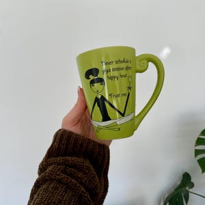 May include: A light green ceramic mug with a whimsical illustration of a woman in a yoga pose. The mug has a curved handle and text that reads, "Never schedule a yoga session after happy hour. Trust me."