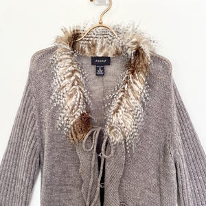May include: A gray knit cardigan with a faux fur collar. The collar features a mix of brown, white, and gray feathers. The sweater has a tie closure and is hanging on a gold hanger.