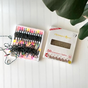 May include: A set of 50 miniature multicolored Christmas lights on a dark green wire, arranged in a white plastic tray. Next to the tray is a white cardboard box labeled Christmas Queen 50 miniature lights for indoor and outdoor use.