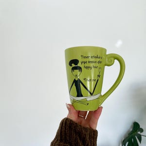 May include: A green ceramic mug with a large handle. The mug features a cartoon illustration of a woman in a yoga pose, with the text "Never schedule a yoga session after happy hour. Trust me."