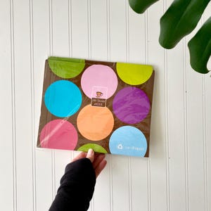 May include: A rectangular package of napkins featuring a brown background with large, colorful polka dots in shades of lime green, pink, bright blue, orange, and purple. The center label displays the text Hot Chocolate JACKIE and cardshaped.