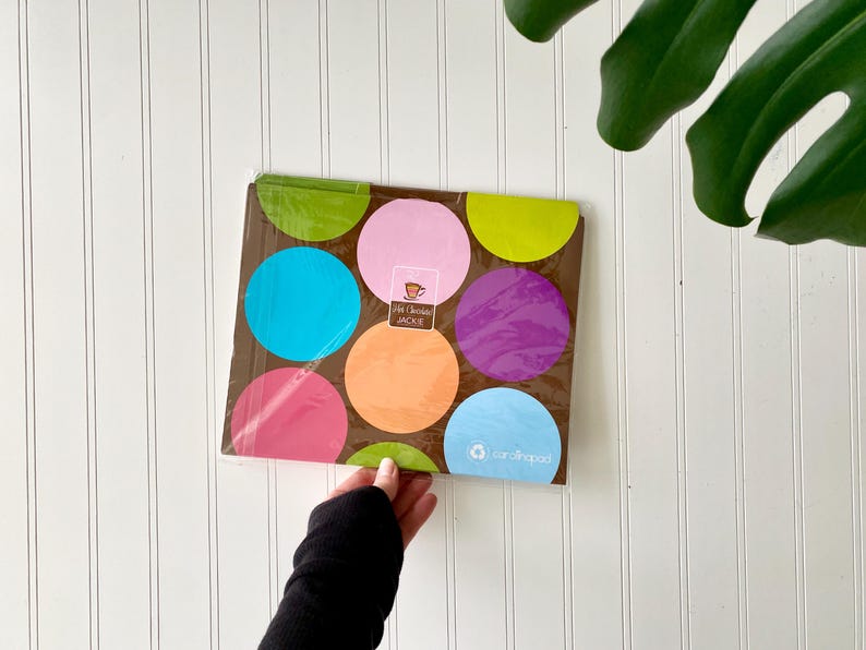 May include: A rectangular package of napkins featuring a brown background with large, colorful polka dots in shades of lime green, pink, bright blue, orange, and purple. The center label displays the text Hot Chocolate JACKIE and cardshaped.