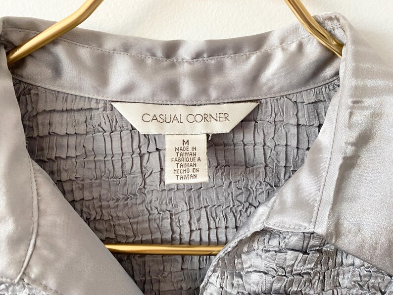 Op de afbeelding: Close-up van een grijs overhemd met kraag en een getextureerd, gerimpeld lijfje. Het overhemd heeft een satijnen kraag en een label met de tekst "Casual Corner" en "Made in Taiwan". Het overhemd hangt aan een gouden hanger.