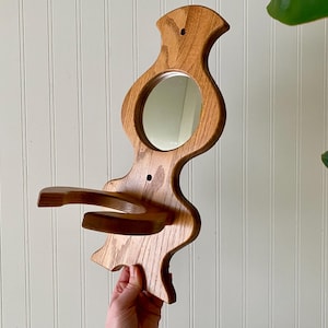 May include: A wooden wall shelf with a mirror and a small shelf. The shelf has a unique, curved design and is made of light brown wood. The mirror is oval-shaped. The shelf is designed to be mounted on a wall.