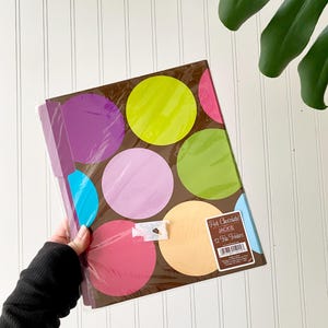 May include: A pack of 12 file folders with a brown background and large, colorful polka dots in shades of lime green, purple, pink, light blue, and peach. A rectangular label on the front reads Hot Chocolate! JACKIE 12 File Folders.