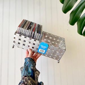 May include: A rectangular silver-toned metal storage basket featuring a woven lattice design and four small spherical feet. The basket contains a row of compact discs and has a blue rectangular product tag attached to the front side with a barcode.