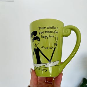 May include: A light green ceramic mug with a whimsical illustration of a woman in a yoga pose, holding a wine glass. The mug has the text "Never schedule a yoga session after happy hour. Trust me."