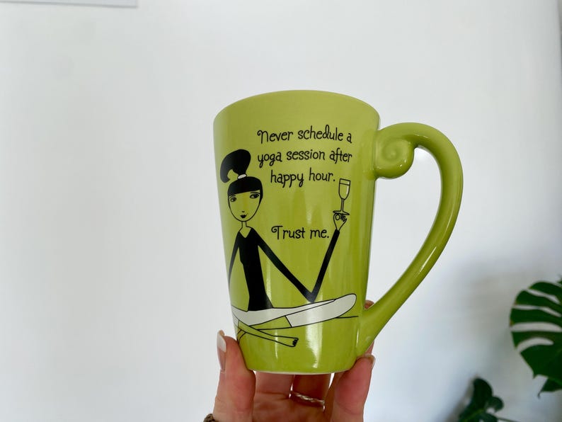 May include: A light green ceramic mug with a whimsical illustration of a woman in a yoga pose, holding a wine glass. The mug has the text "Never schedule a yoga session after happy hour. Trust me."