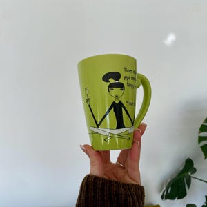 May include: A light green ceramic mug with a black handle. The mug features a cartoon illustration of a woman in a yoga pose holding a wine glass, with the text "Never skip yoga, happy hour, or Friday".